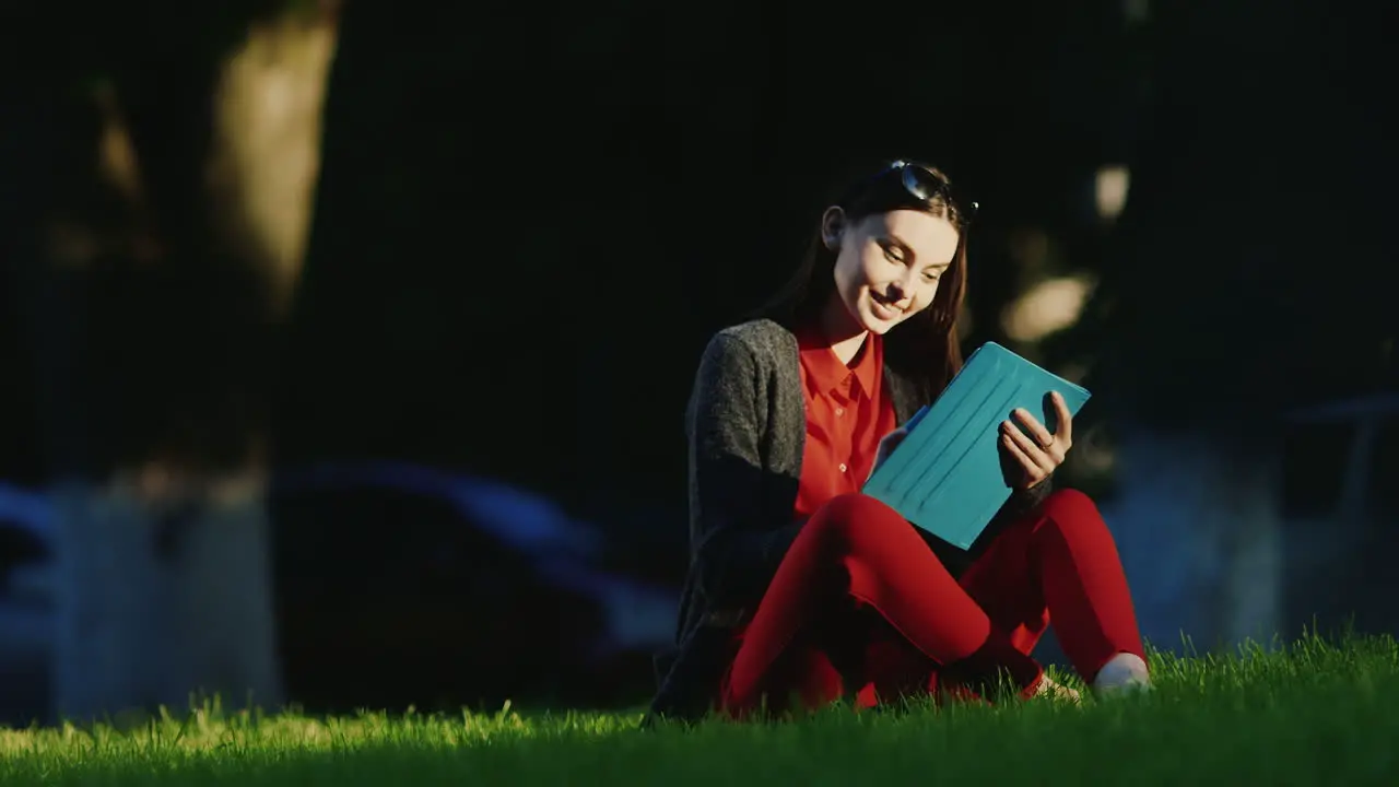 Young Woman Enjoys The Tablet In The Park Sitting On The Grass His Face Lit By The Evening Sun Hd Vi