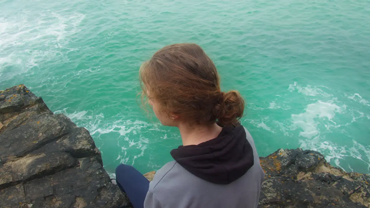 Caucasian Teenage Boy Looking Away With A Sad Face While Sitting on Rocky Cliff Over The Ocean Overhead Zoom out