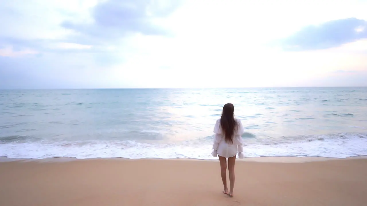 A young woman standing on the beach with her back to the camera watches the surf roll in