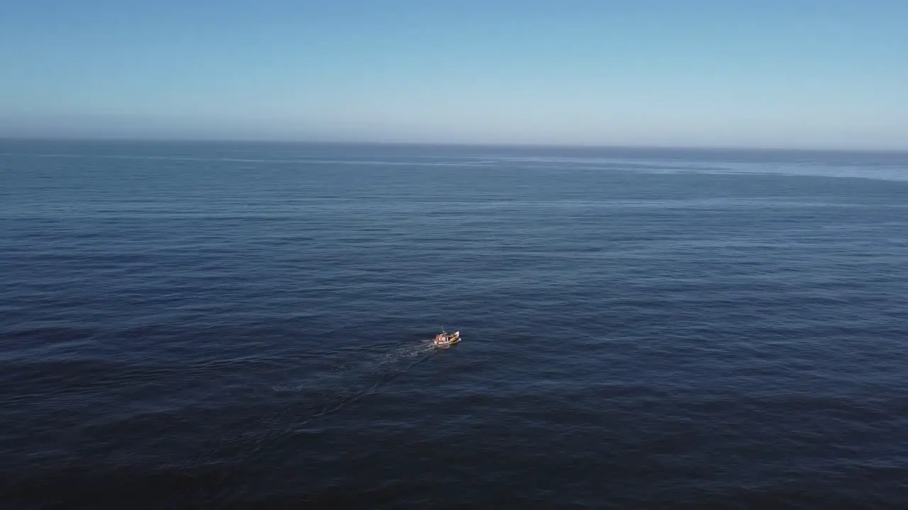 Aerial view of a small white boat sailing in an immense sea getting further and further from the coast