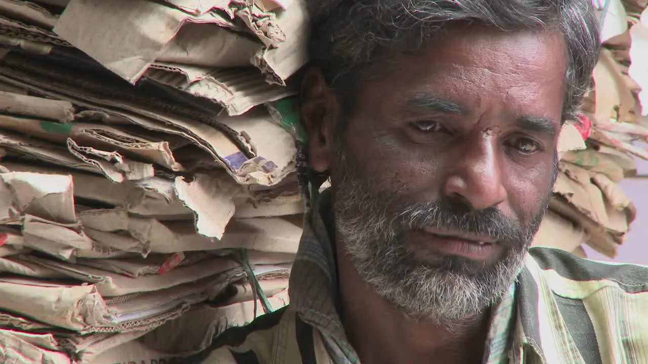 Older bearded man leans up against a tall stack of flattened cardboard boxes