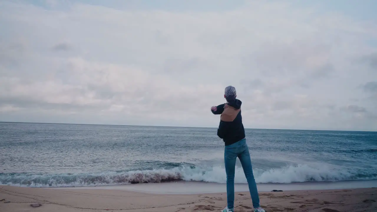 Young Tall Guy With Jacket Throwing Pebbles and Rocks Into The Ocean Static Wide Shot