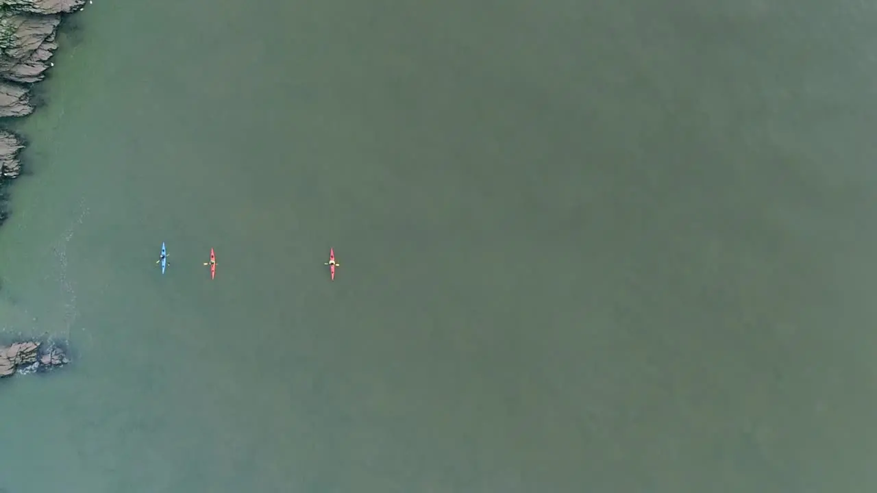 Top down aerial of three kayakers swimming along a rocky shore