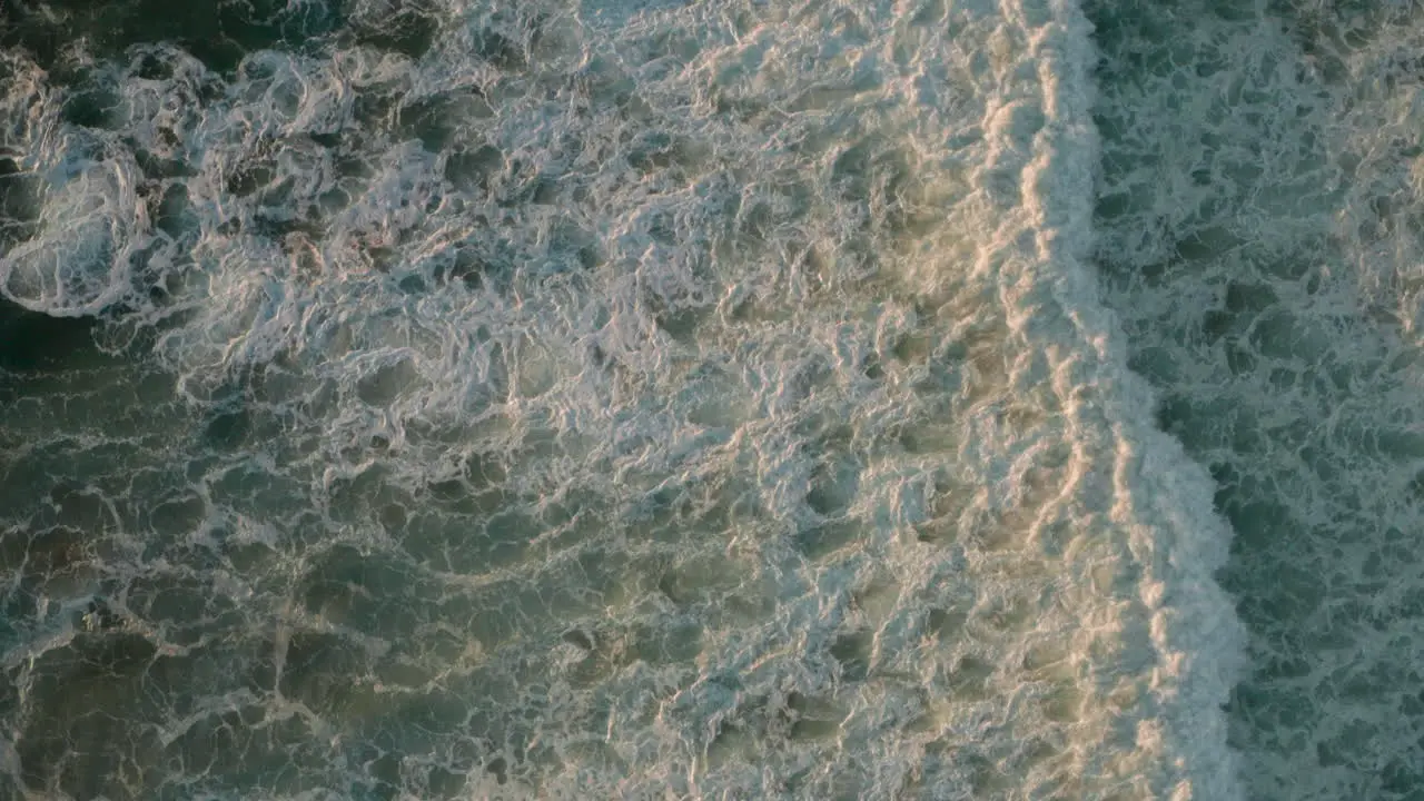 Top down aerial shot over white foaming waves in a blue sea at golden hour