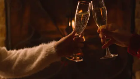 Close Up of Two People Bringing Their Champagne Glasses Together In Front of Burning Fireplace