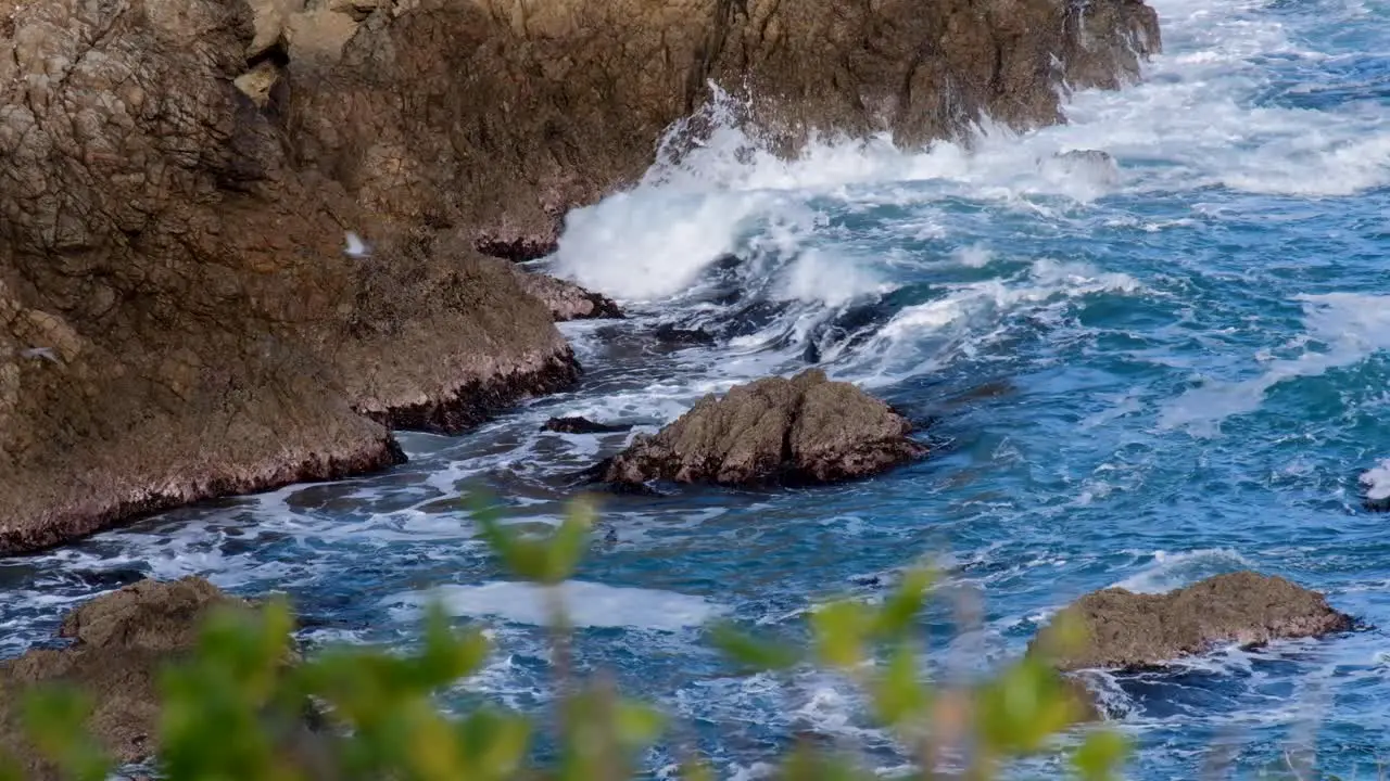 A flock of seagulls flying over rough choppy sea white wash waves smashing against a rocky cliff of ocean coastline