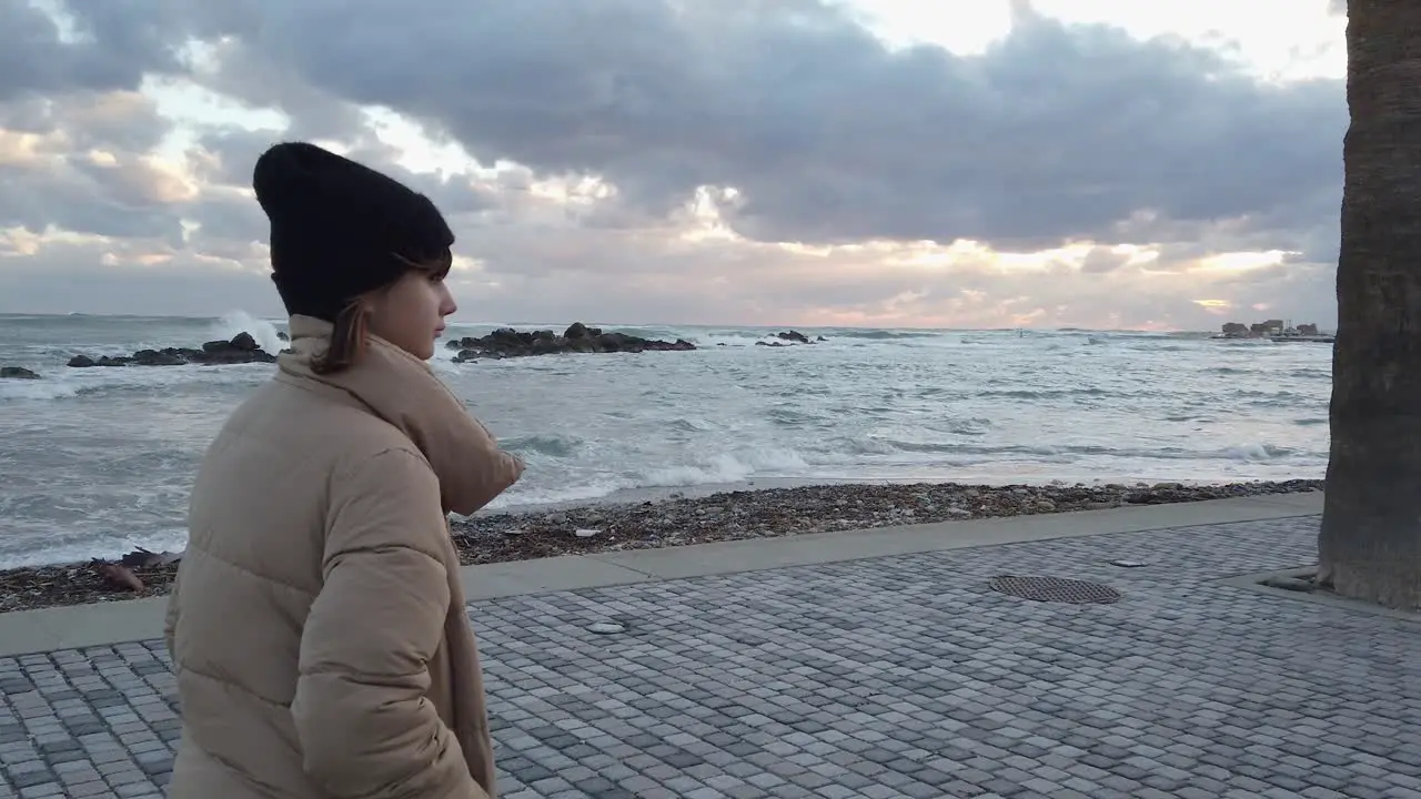 A young girl walks alone along the boardwalk with angry sea waves crashing ashore in the background in slow motion