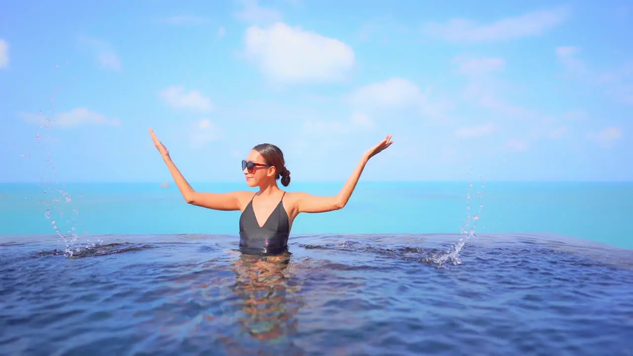 Happy Asian woman in black bathing suit and red sunglasses inside rooftop infinity pool water splashing water with hands cloudy sky and sea on background