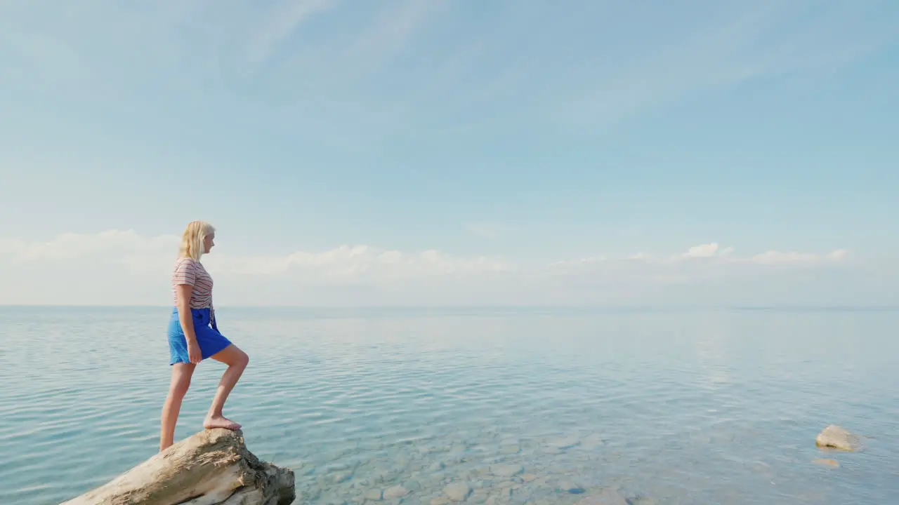 Active Woman Traveler Stands On The Edge Of A Log Admires A Beautiful View Of The Calm Sea And Blue 