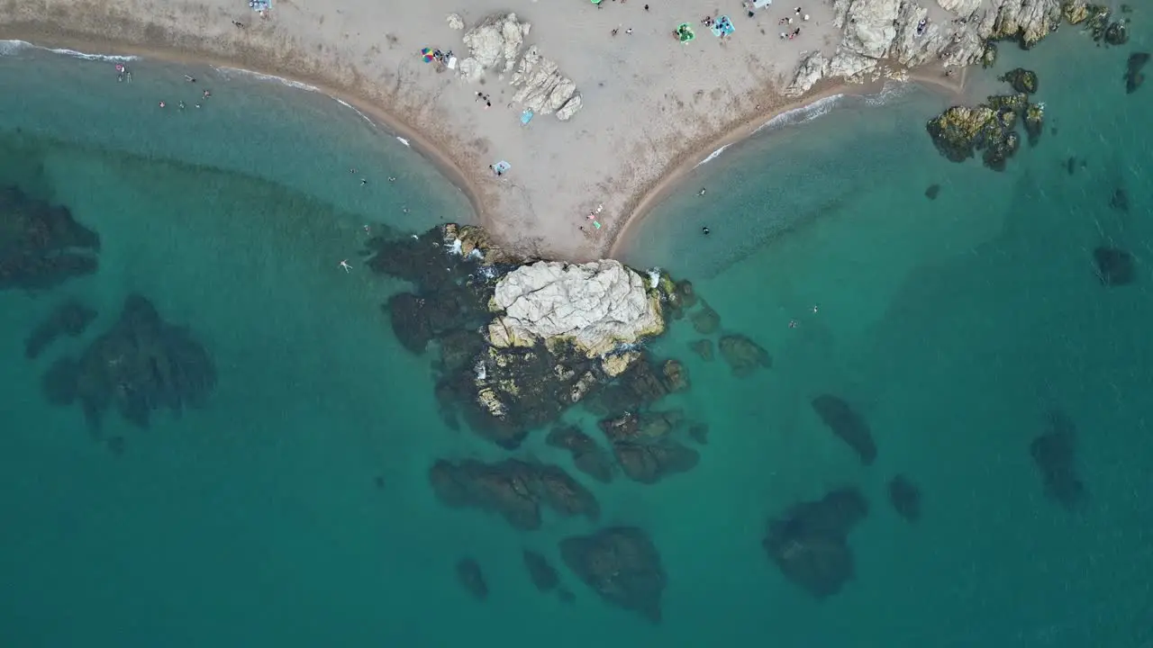 zoomed top view of a rock in the middle of two beaches with people bathing and waves crashing on the shore