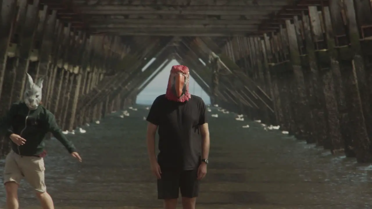 Two masked people dance under the legs of a pier