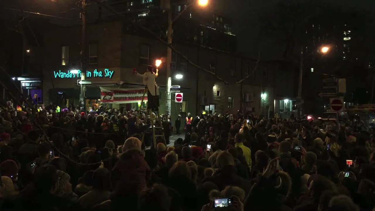 Wide shot of a night time gathering in Toronto's Kensington Market as fire performer on a high ladder breathes fire to mark the beginning of the Winter Solstice