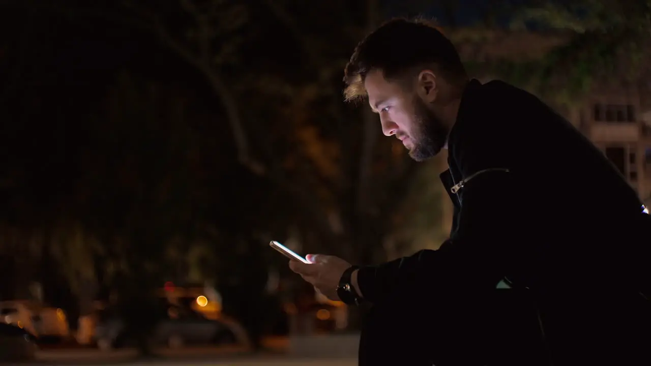 Night time medium shot of a man sitting on a bench in a park scrolling-searching on a smartphone