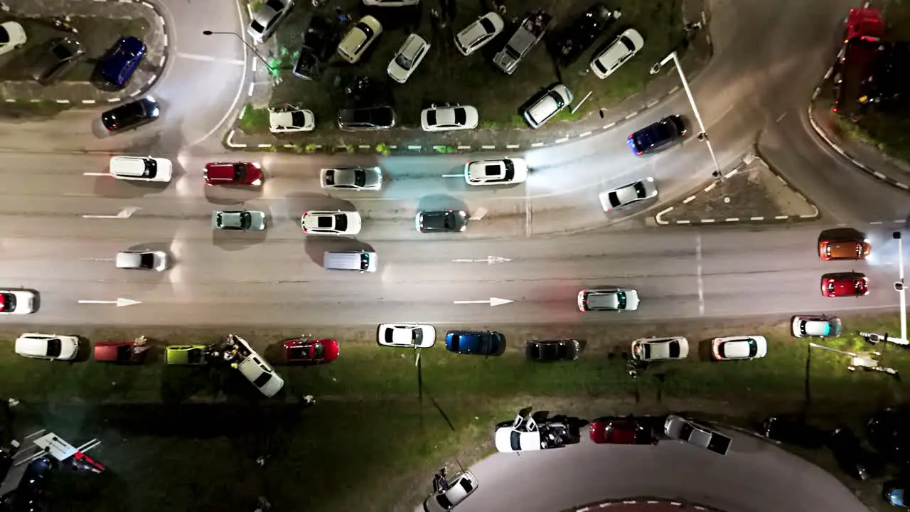 Drone ascends above cars parked on grass median and turning onto on ramp at night