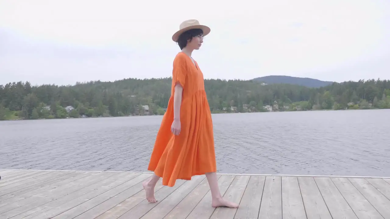 Pretty Girl in Vibrant Orange Dress Walking on Lakeside Wooden Deck