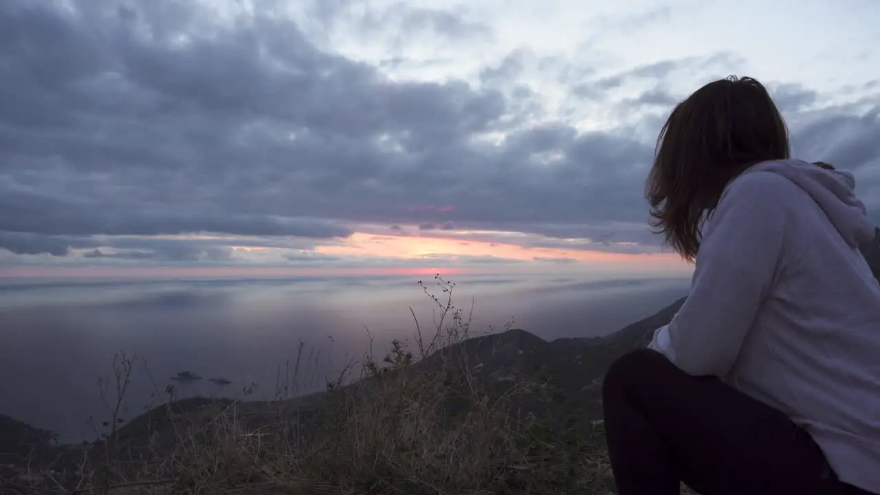 Girl squat on a high cliff and watch the sunset through cloudy sky over Adriatic sea-1