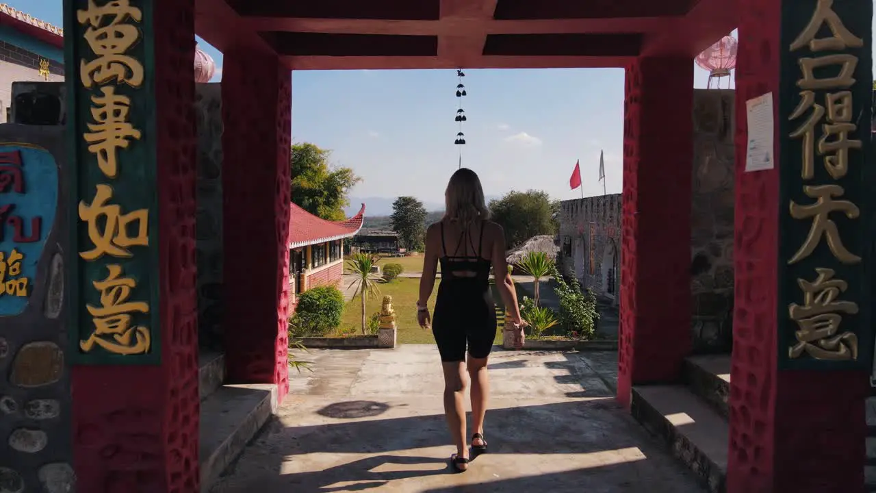 Blonde woman in black sportswear walks through the gate of the historic Chinese village of Santichon in Thailand on a warm summer afternoon