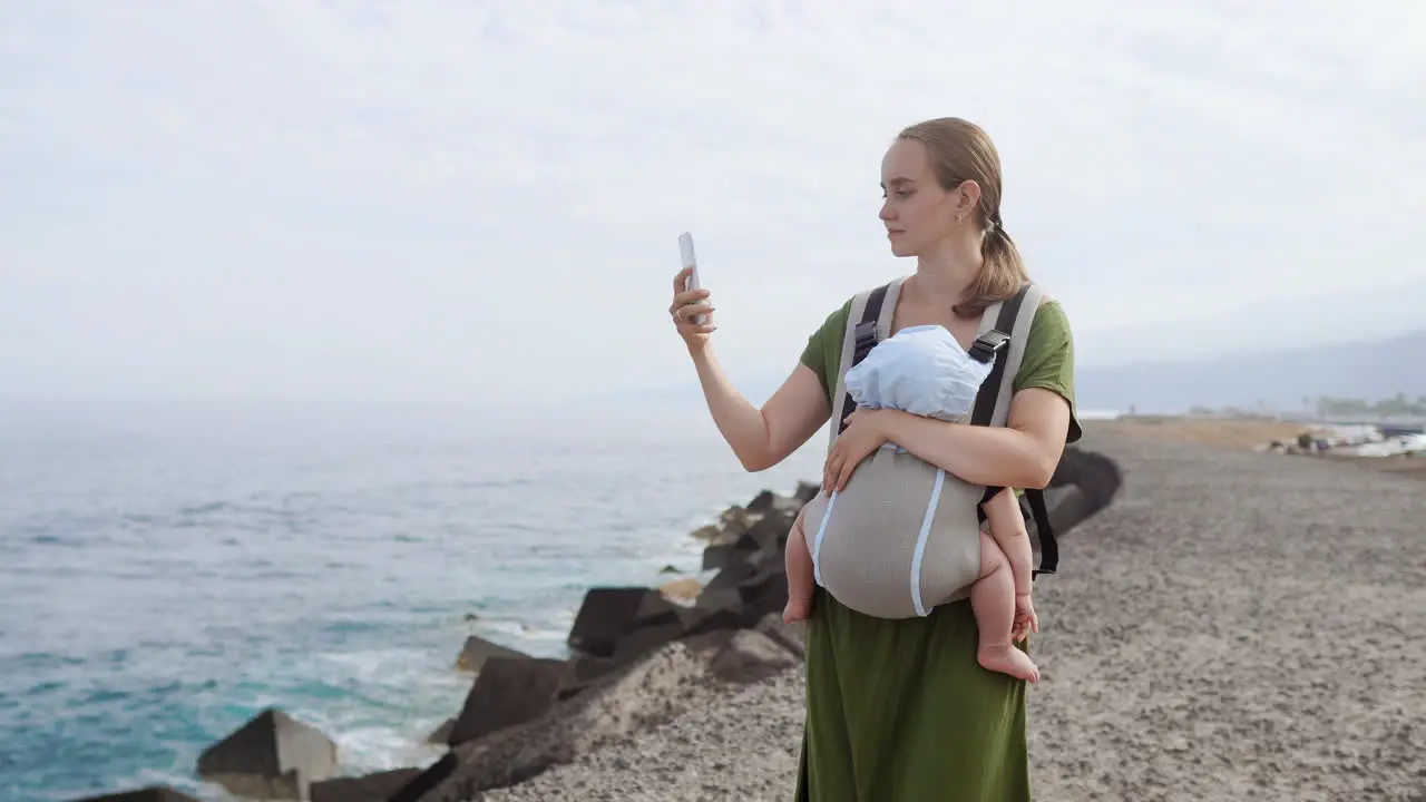 Against the backdrop of the ocean a mother and her baby spend a delightful summer day outdoors The child is cozily nestled in a kangaroo backpack as they walk along