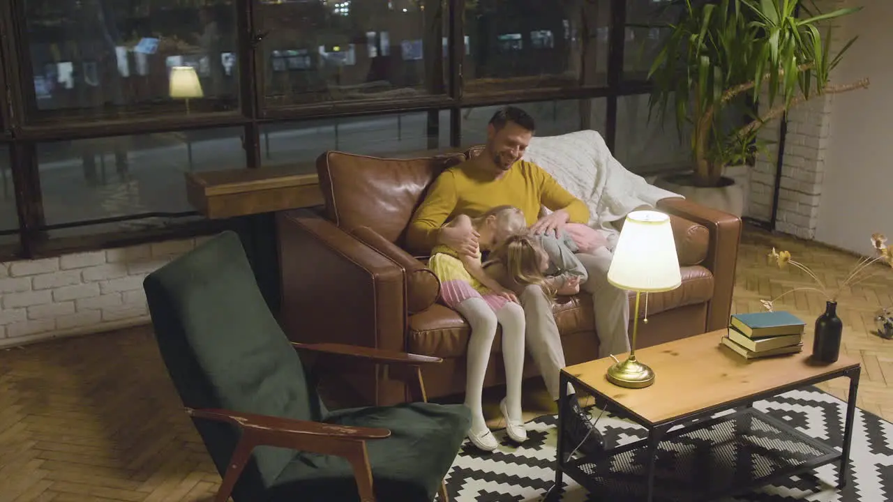 Happy Father Sitting On Sofa With His Two Sleepy Little Girls Who Leaning Their Heads On His Lap 1