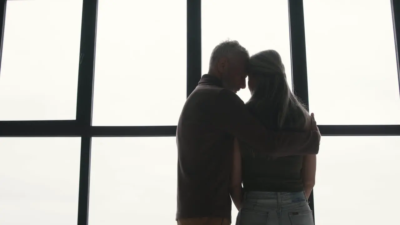 Back View Of Middle Aged Man Embracing His Sad Wife And Trying To Comfort Her While Standing Together At Window At Home