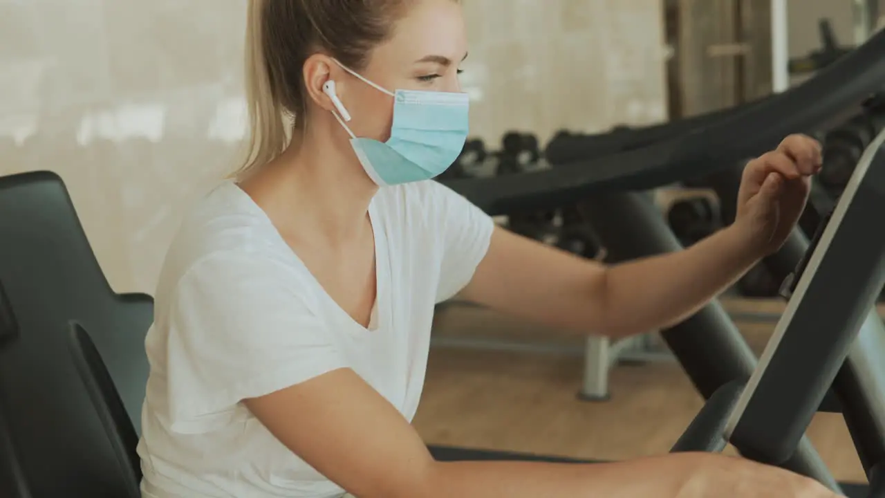 Young Athlete Female With Face Mask Uses A Smartphone And An Exercise Machine In The Gym