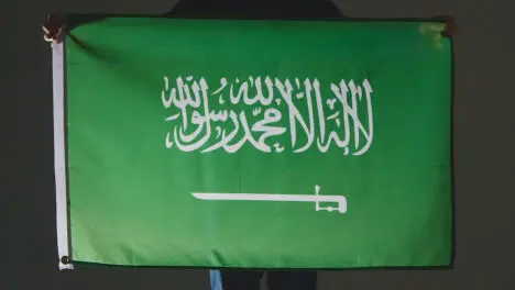 Studio Shot Of Anonymous Person Or Sports Fan Holding Flag Of Saudi Arabia Against Black Background