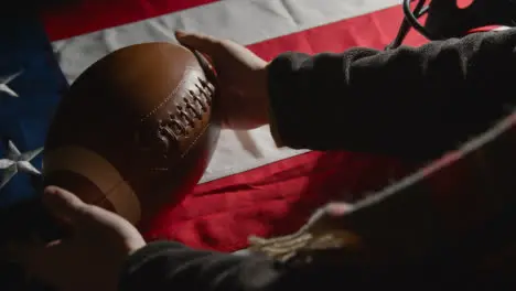 Studio Shot Of Person Picking Up American Football With Helmet On Stars And Stripes Flag 