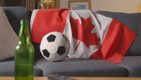 Flag Of Canada Draped Over Sofa At Home With Football Ready For Match On TV