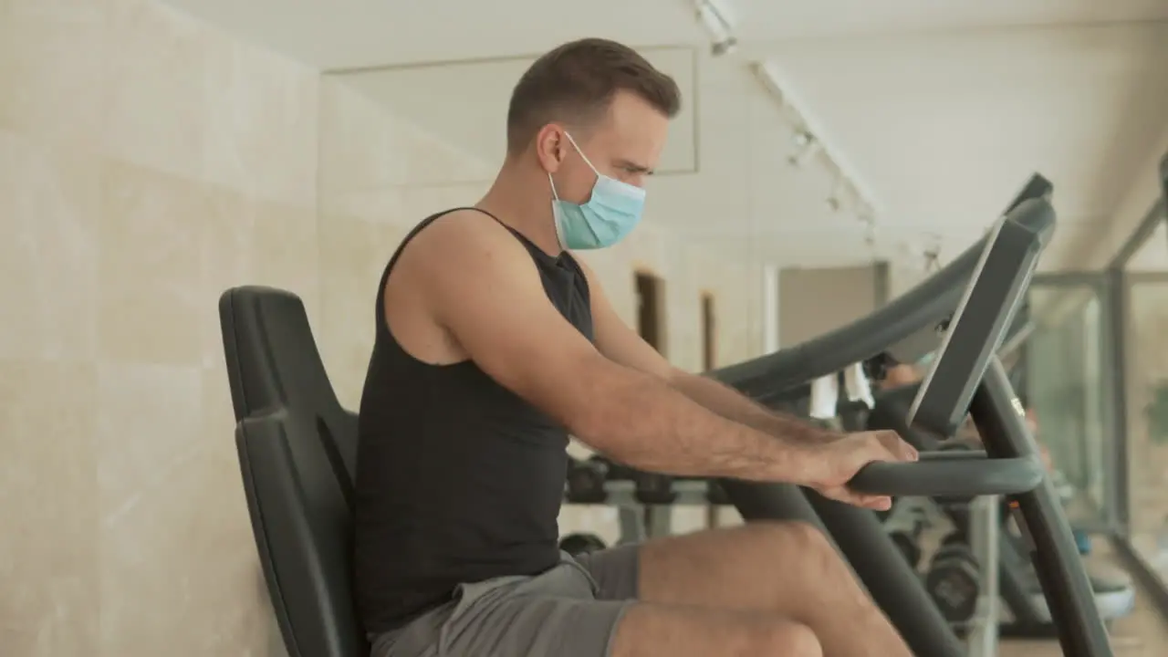 Strong Male With Face Mask Uses An Exercise Machine And Drinks Water In The Gym