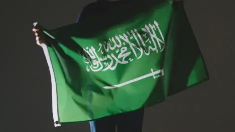 Studio Shot Of Anonymous Person Or Sports Fan Waving Flag Of Saudi Arabia Against Black Background