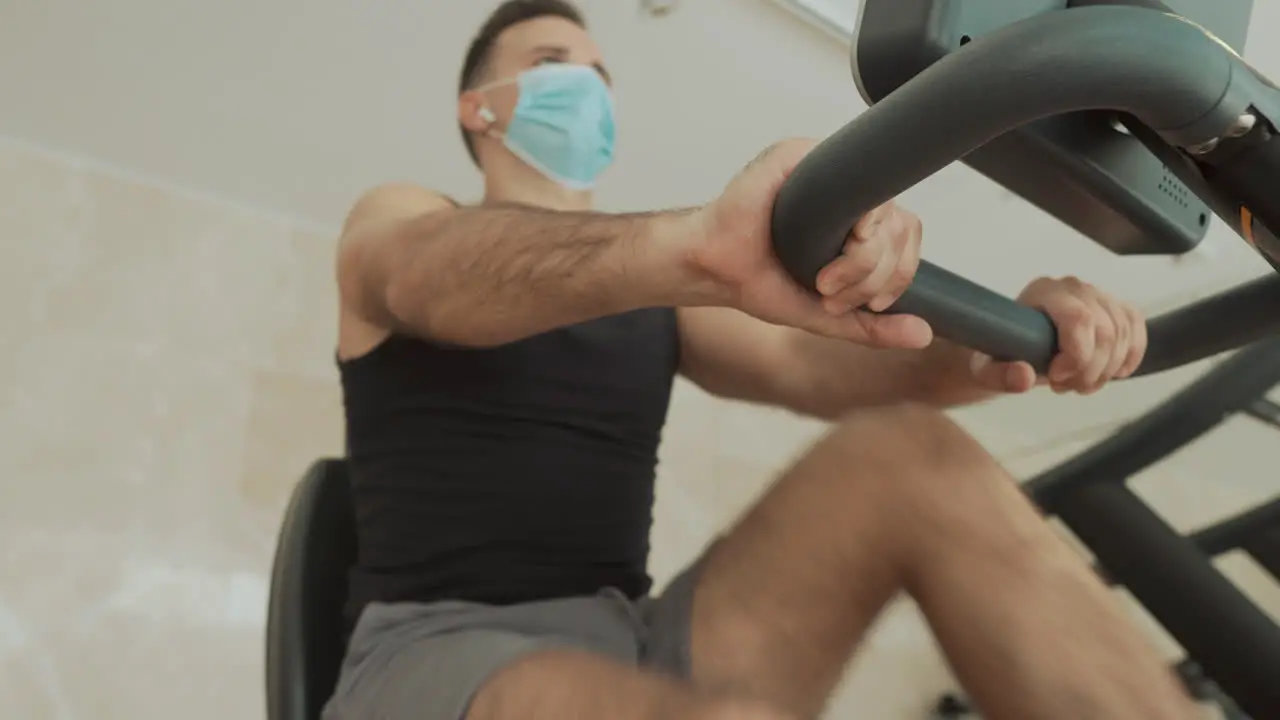 Young Strong Athlete Male Man With Face Mask Uses An Exercise Machine And Drinks Water In The Gym