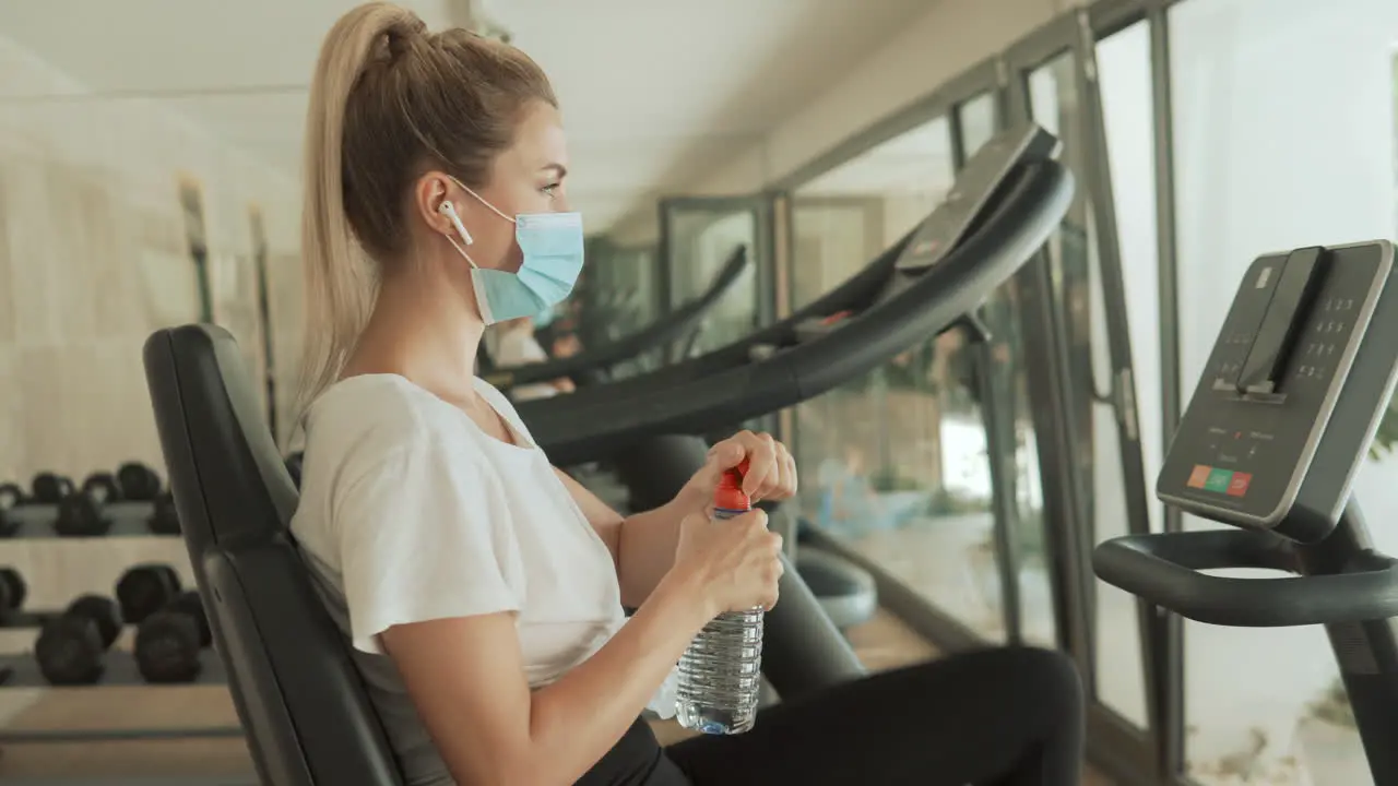 Young Athlete Female With Face Mask Uses An Exercise Machine And Drinks Water In The Gym