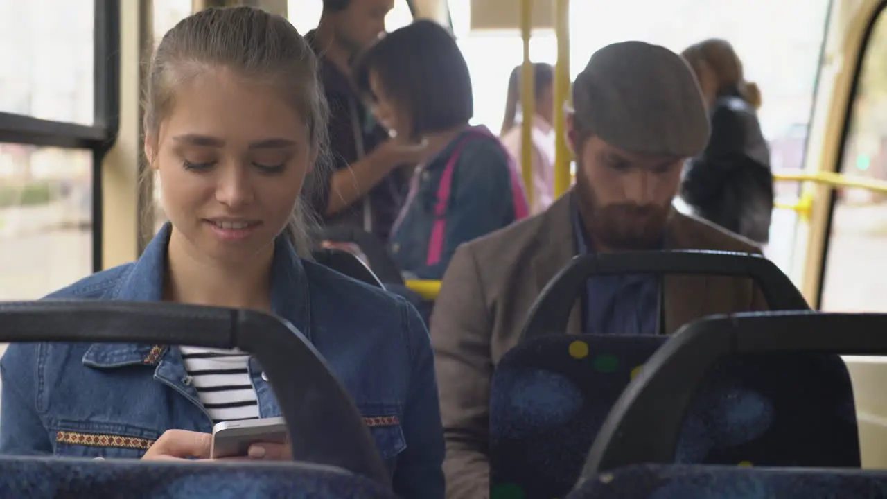 Young Woman Sitting On A Bus While Using Smartphone Behind Is A Man In A Beret Looking At Smartphone And People Standing