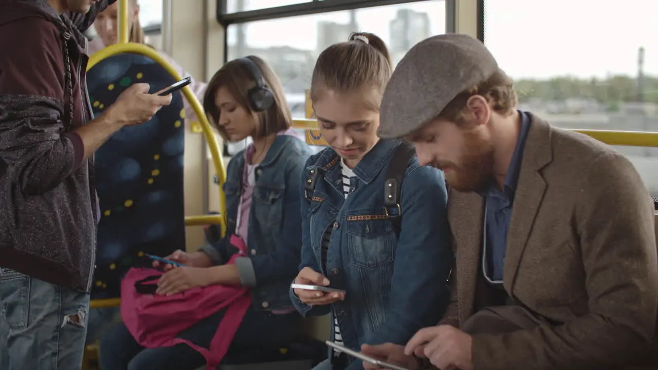 Young Man With Beard And Beret Using Smartphone While Talking To Young Girl Holding Smartphone Both Traveling By Bus In The City