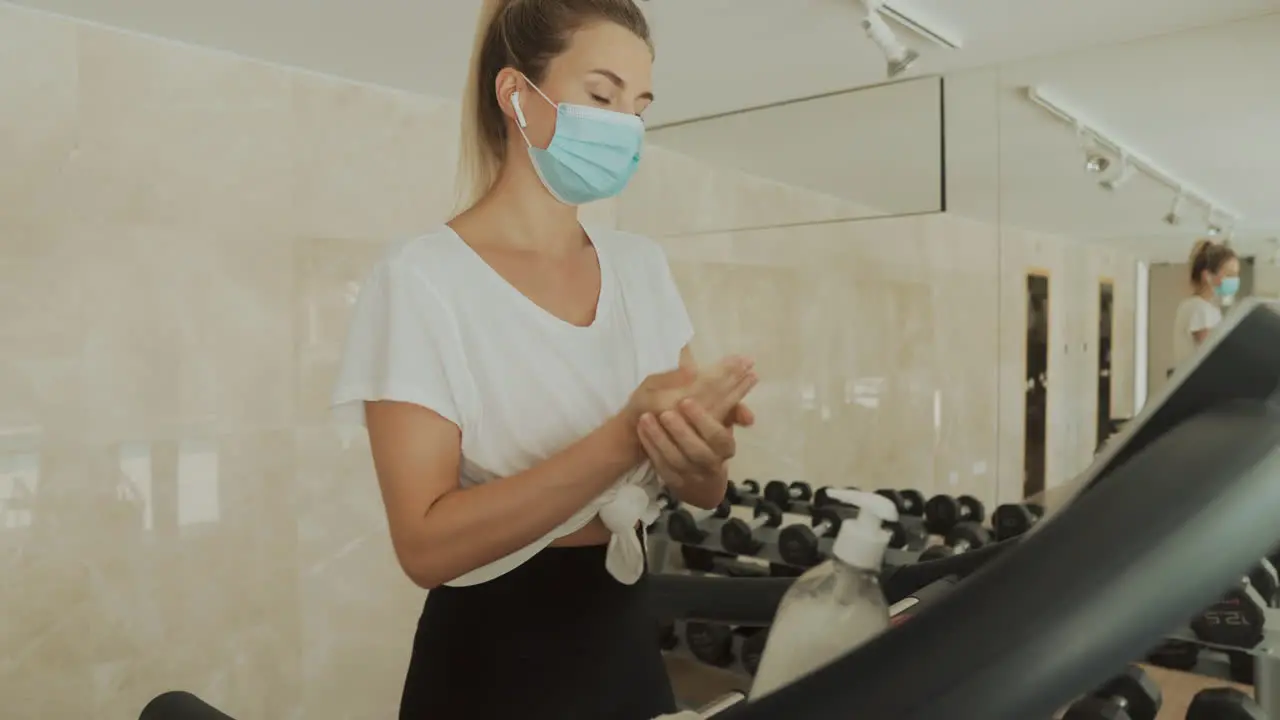 Young Athlete Woman With Face Mask Uses An Exercise Machine And Hand Sanitizer In The Gym