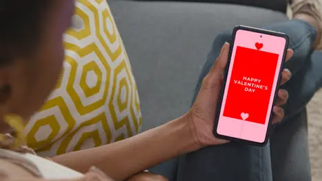 Close Up Of Woman Sitting On Sofa At Home Looking At Happy Valentine's Day Message On Mobile Phone