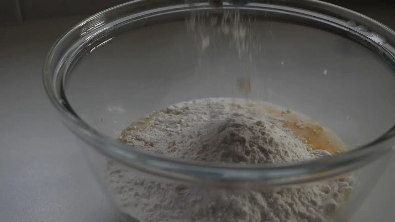 Person sprinkles flour from blue measuring cup into egg baking mixture in glass bowl