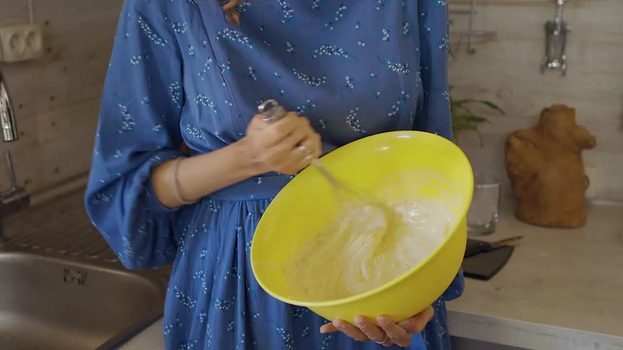 Wide shot of a woman in a blue dress mixing cake batter in a yellow large bowl