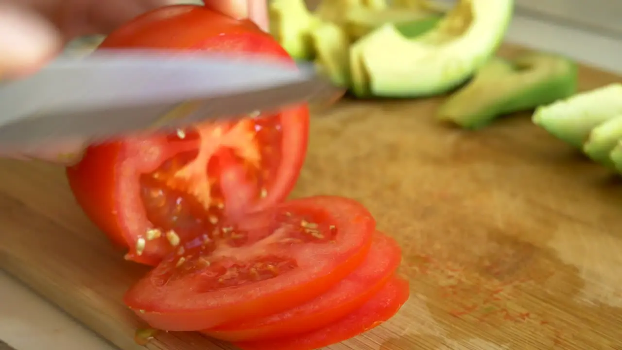 A chef cut and slices a ripe red tomato and green avocado vegetables on a bamboo cutting board while making a healthy vegan meal in kitchen