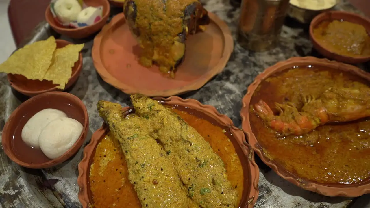 Profile view of fishes rasgullas prawns papad chutney and some other dishes given on plates made up of clay and water on bronze glass