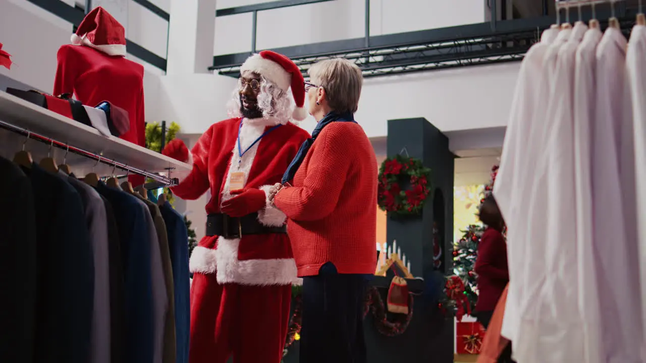 Employee dressed as Santa Claus presenting materials and price informations about red garment piece to elderly client looking to buy gift for family member in Christmas decorated clothing store