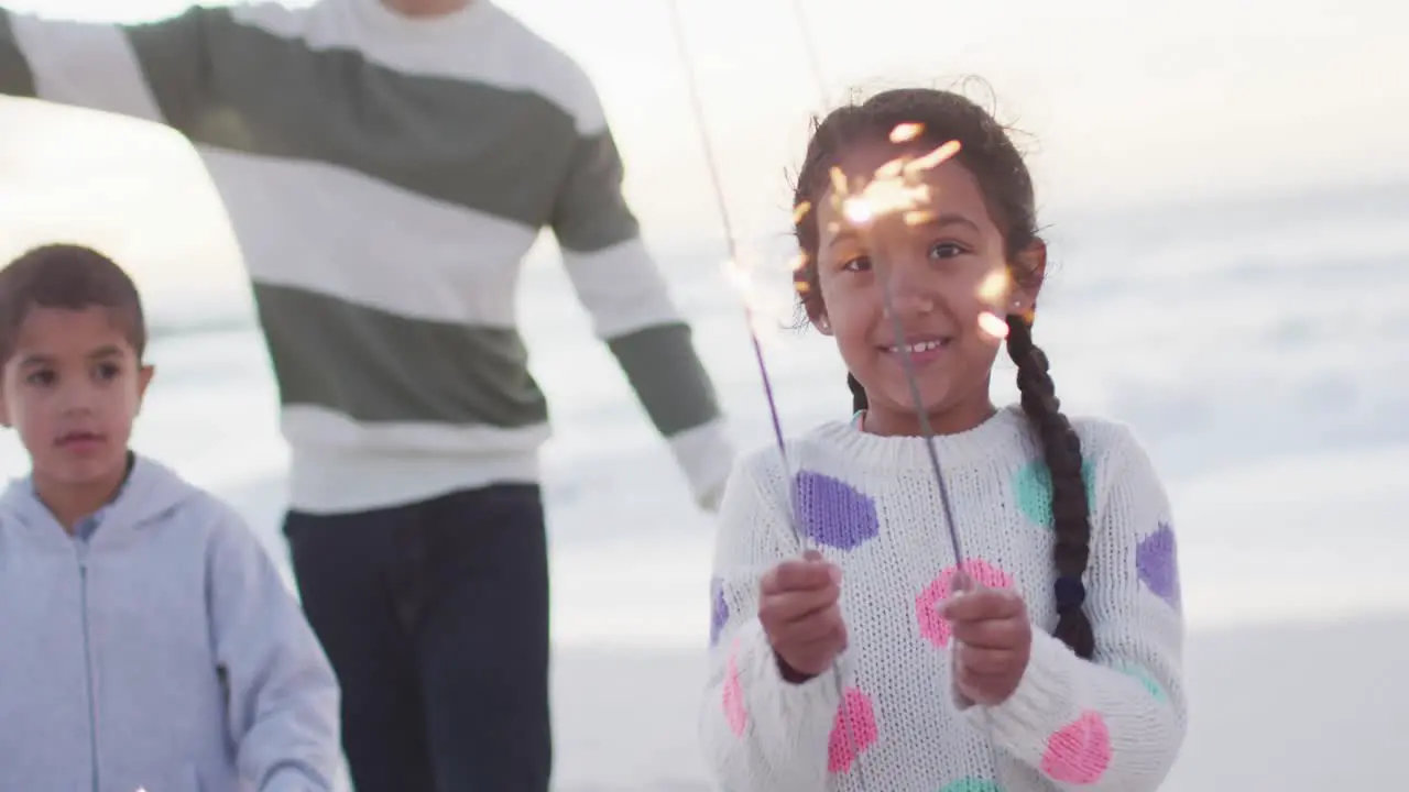 Portrait of happy hispanic girl playing with sparklers on beach with family at sunset