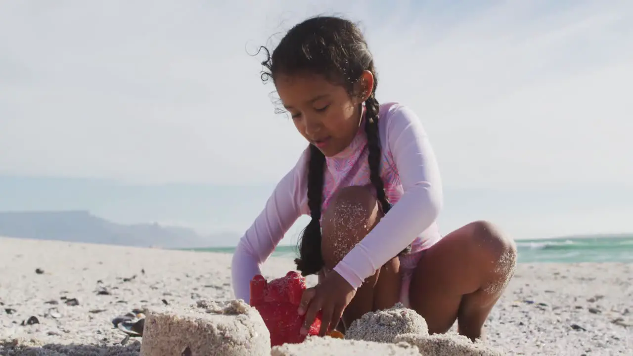 Happy hispanic girl sitting on beach and building sand castle