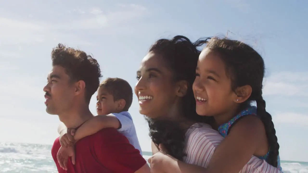 Happy hispanic parents carrying children on piggyback and walking on beach