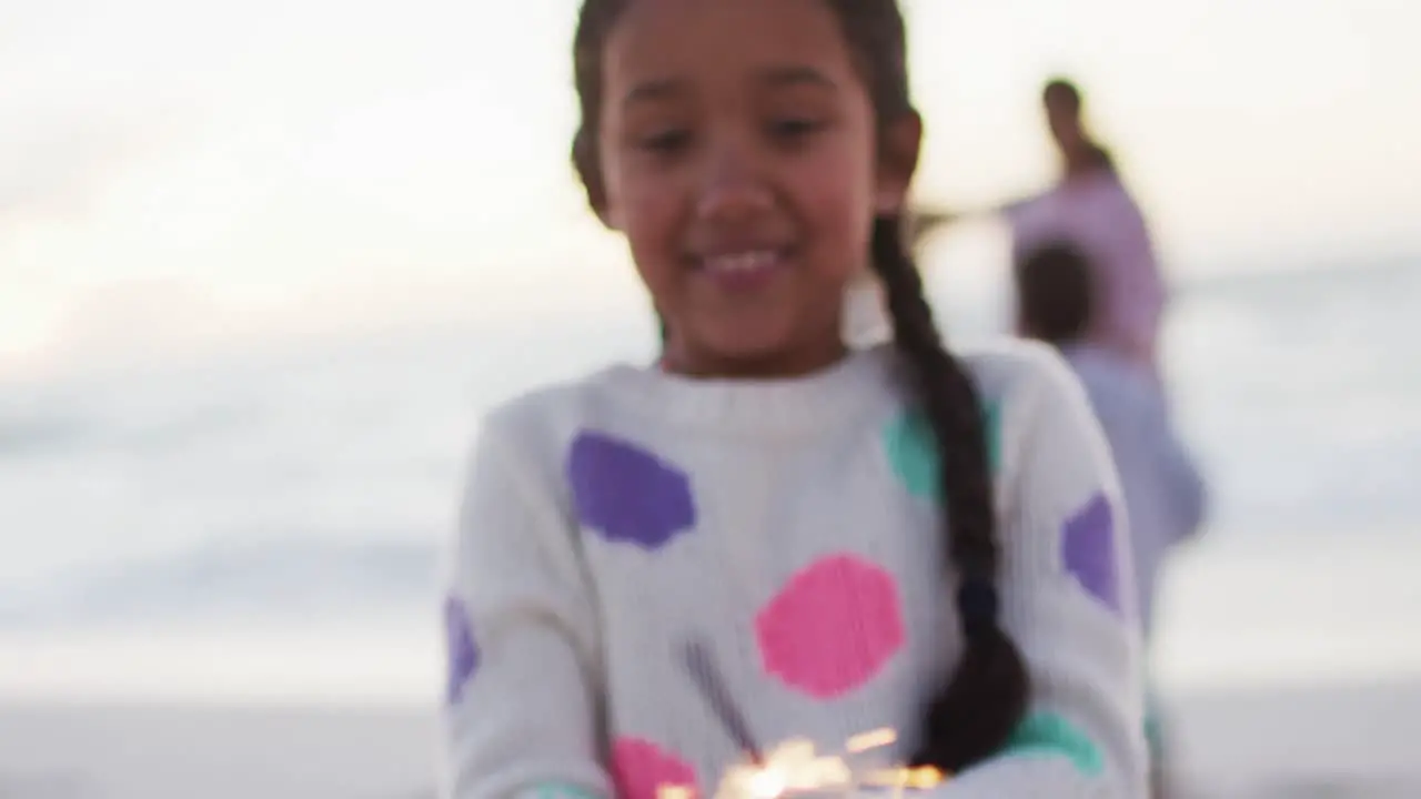 Portrait of happy hispanic girl playing with sparklers on beach at sunset