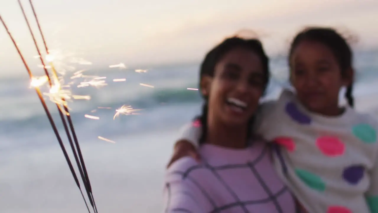 Portrait of happy hispanic mother and daughter playing with sparklers on beach at sunset