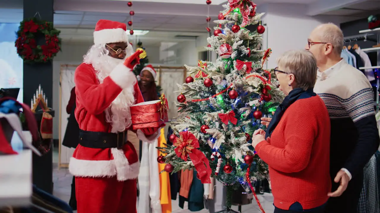 Employee in xmas adorn fashion boutique wearing Santa Claus costume and customers gathered around beautifully decorated Christmas tree in clothing store during winter holiday season