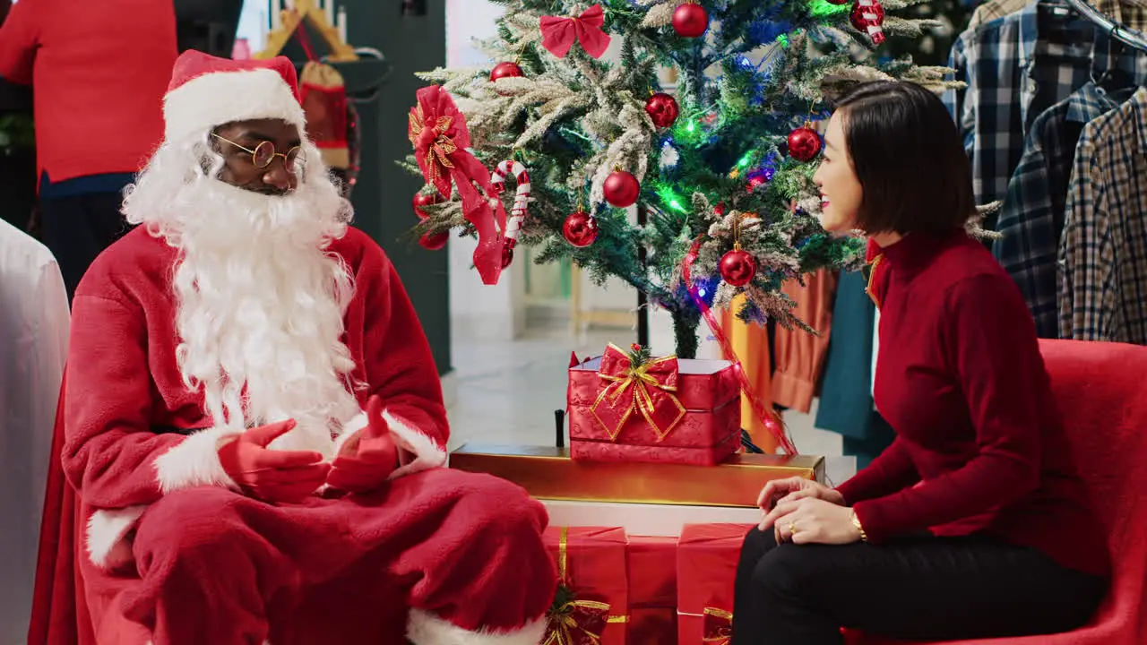 Asian woman engaging in conversation with employee impersonating Santa Claus during festive Christmas shopping experience Customer receiving xmas present from worker in fashion shop