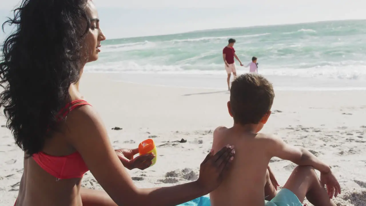 Back view of hispanic mother applying sun cream on son back on beach