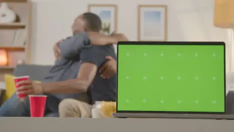 Two Men Celebrating Watching Sports Game On TV At Home With Green Screen Laptop In Foreground 